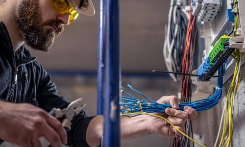 A male electrician works in a switchboard with an electrical connecting cable, connects the equipment with tools.